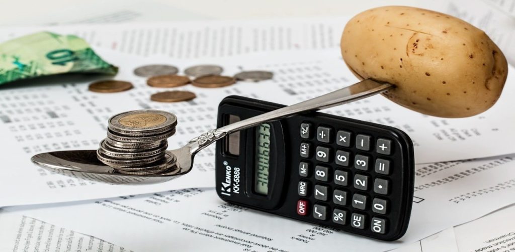 Calculator flipped horizontally on its side with a spoon being balanced with a potato on the handle and coins on the other end